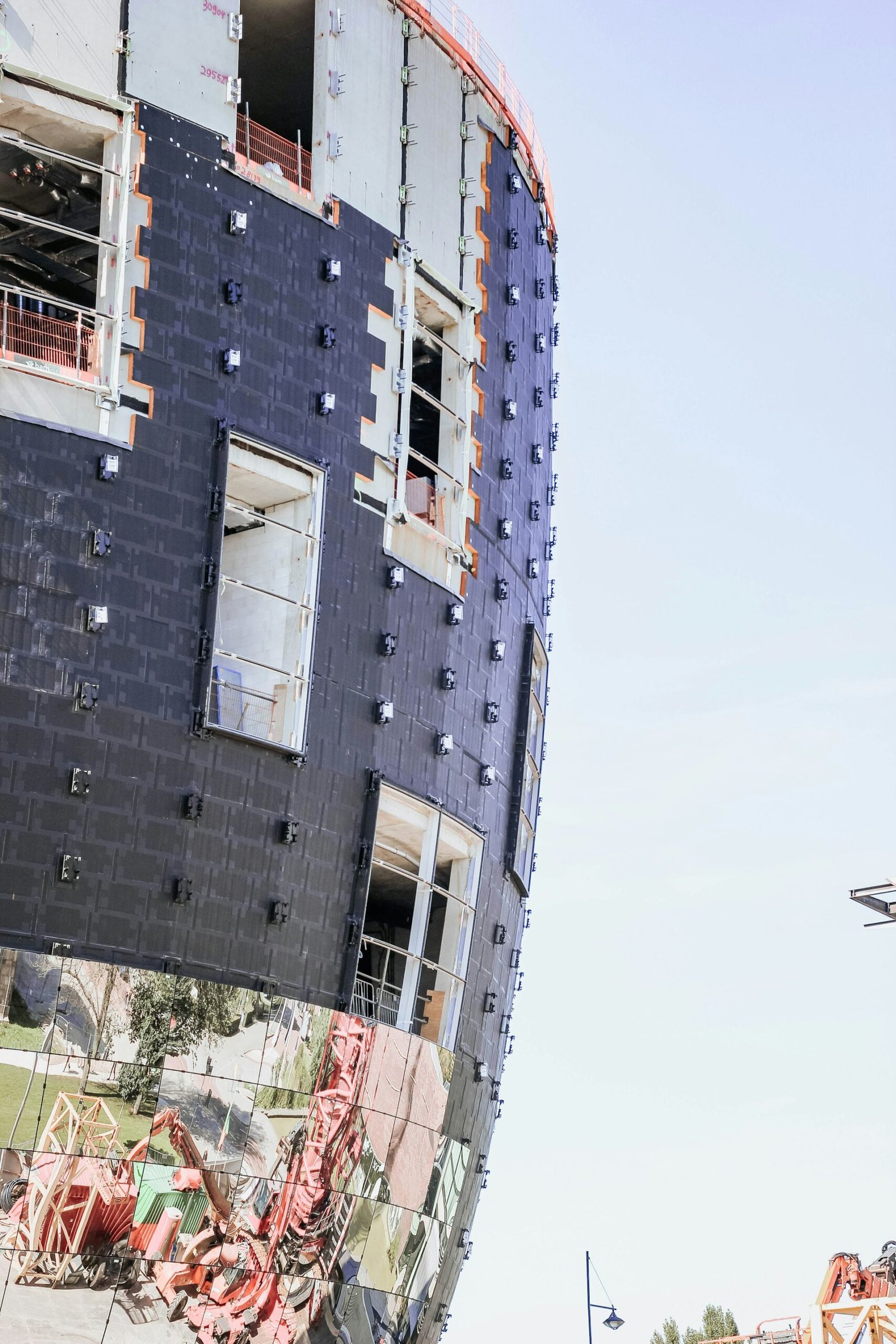 A modern building under construction with a curved facade is wrapped in black materials and features reflective lower panels. Openings for windows and evidence of construction materials are visible, with scaffolding and a crane appearing in the surroundings.